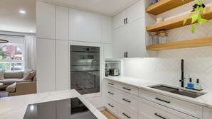 Uma cozinha moderna com armários brancos, forno embutido, máquina de café e uma prateleira de madeira com potes.