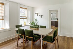 Ein Essbereich mit einem weißen Tisch, grünen Stühlen, einer Pflanze als Mittelstück und einer Küche im Hintergrund.