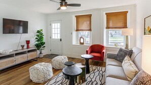 A living room with a flat-screen TV, a red chair, a sofa, ottomans, a ceiling fan, and a window with blinds. - 4 Mi to Cedar Point! Spacious Home w/ Fenced Yard (Sandusky)