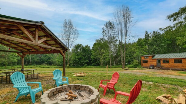 A fire pit with chairs, a covered picnic table, and a cabin in a wooded area. - Catskills Airstream! Pavilion & Fire Pit Access (Franklin)