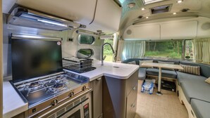 A compact kitchen with a gas stove, a microwave, and a small dining area with a table and seating. - Catskills Airstream! Pavilion & Fire Pit Access (Franklin)