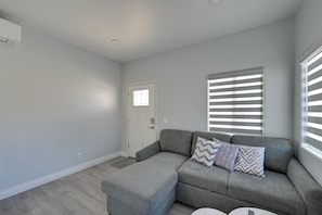 Ein modernes Wohnzimmer mit einem grauen Ecksofa, einer weißen Tür und einem Fenster mit horizontalen Jalousien.