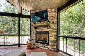 Ein Kamin aus Stein mit darüber montiertem Fernseher, ein Feuer brennt darin, und eine Terrasse mit Gartenmöbeln.