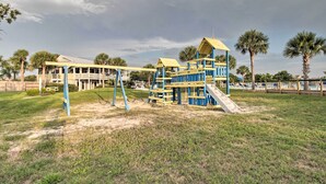 Un parque infantil con tobogán y columpios, rodeado de césped y palmeras.