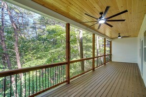 Une terrasse en bois couverte, dotée d’un ventilateur de plafond, qui offre une vue sur une zone boisée.
