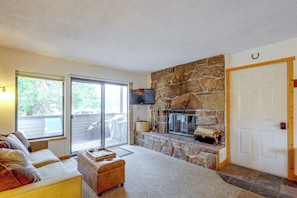 A living room with a stone fireplace, a flat-screen TV, a sofa, and a sliding glass door leading to an outdoor area. - Fraser Condo w/ Pool & Hot Tubs: Close to Trails! (Fraser)