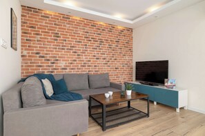 A modern living room with a brick wall, grey sofa, coffee table, TV, and a small plant. - Family Apartment with Balcony | Underground Parking (Kraków)