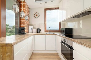 A modern kitchen with white cabinets, a brick accent wall, a large window, and a view of the city. - Family Apartment with Balcony | Underground Parking (Kraków)