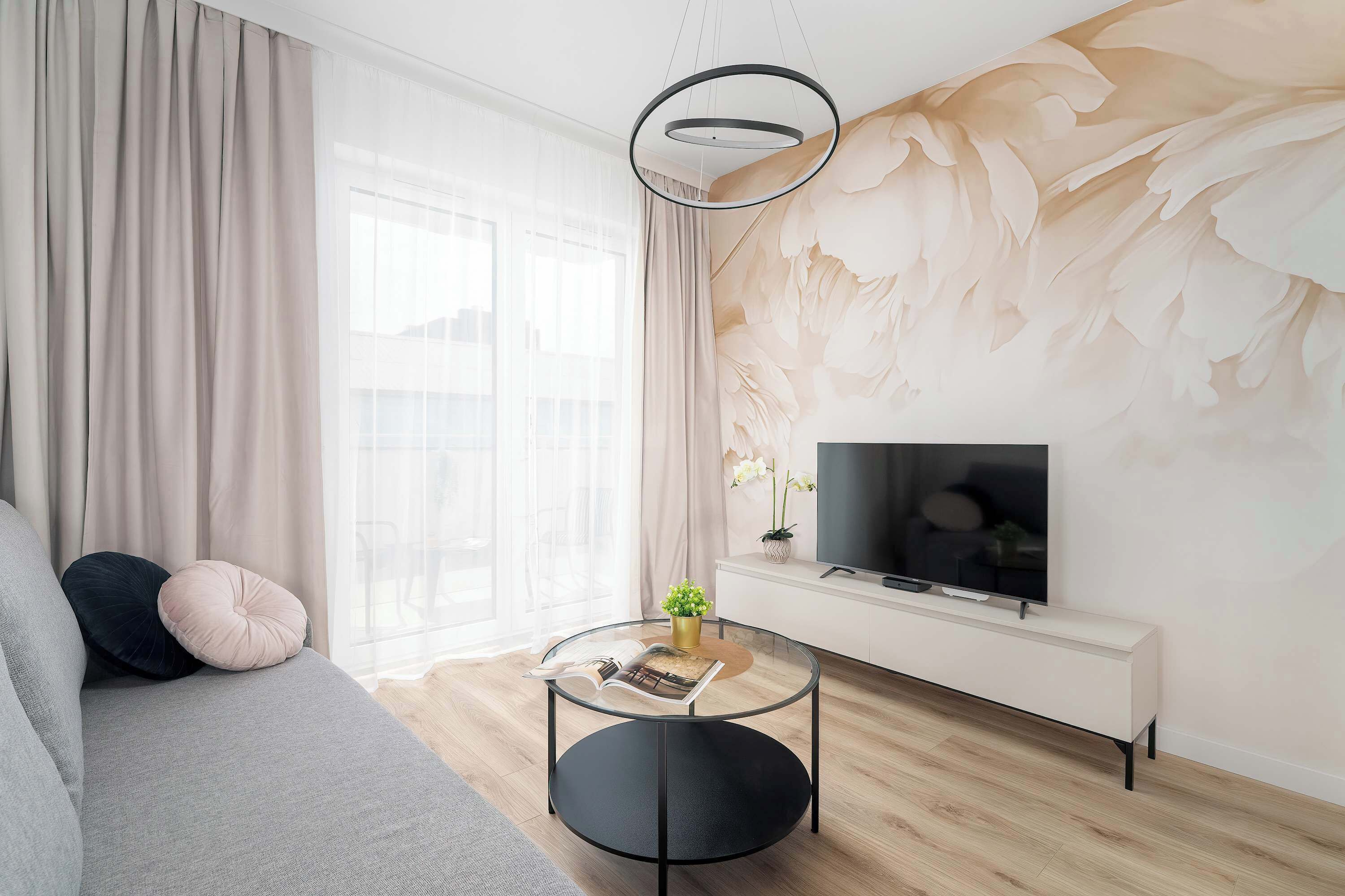 En moderne stue med sofa, rundt sofabord, TV på hvidt skab og stort vindue med lette gardiner.