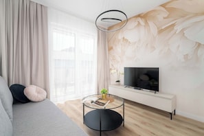 A modern living room with a sofa, a round coffee table, a TV on a white cabinet, and a large window with sheer curtains. - Cozy Apartment with Balcony | Underground Parking (Poznań)