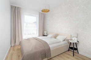 A modern bedroom with a large bed, a bedside table with a lamp, and a window with curtains. - Cozy Apartment with Balcony | Underground Parking (Poznań)