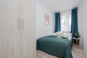 Een slaapkamer met een bed, nachtkastje, lamp, potplant en een raam met gordijnen.