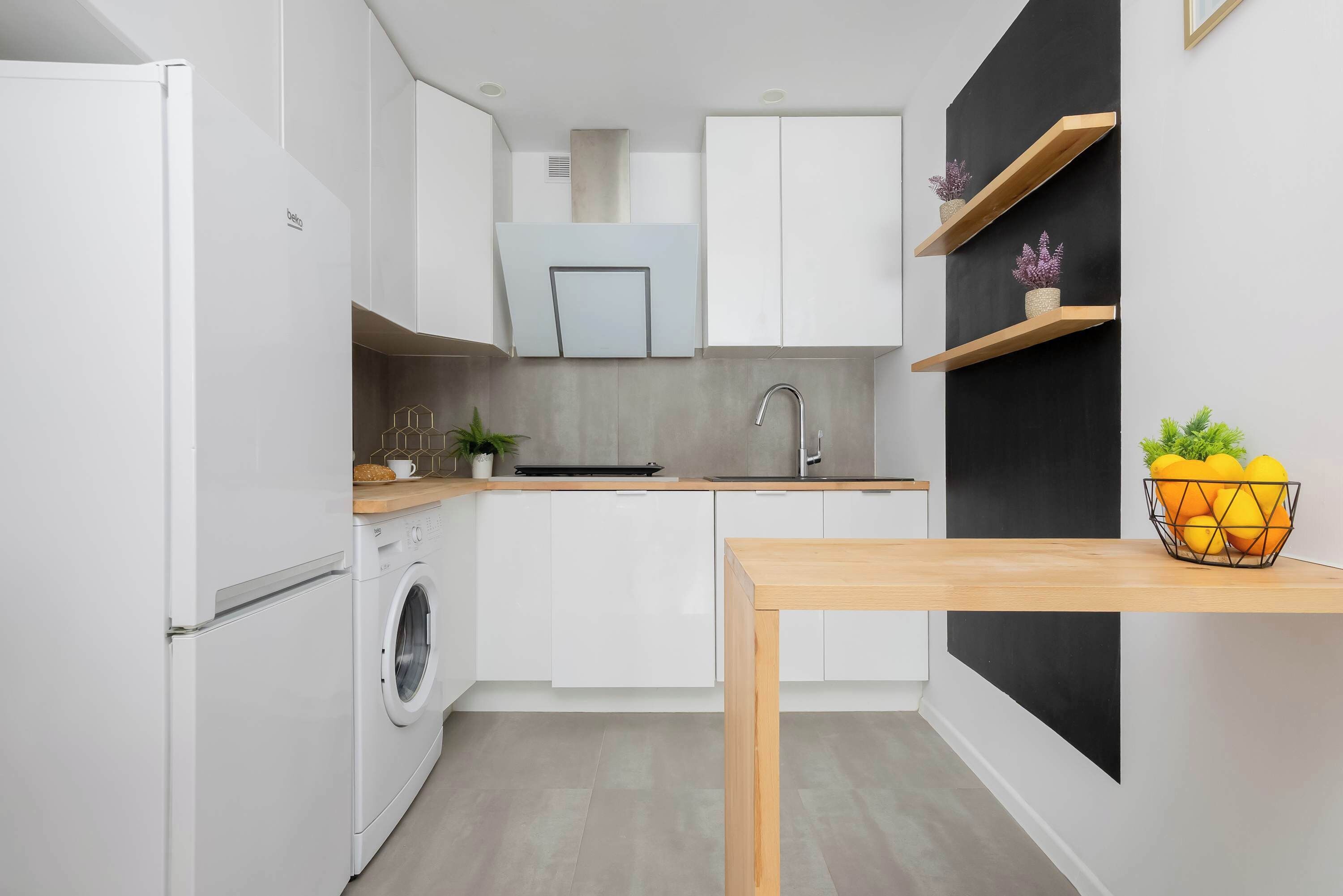 A compact kitchen with a washing machine, a sink, and a wooden table with a fruit bowl.