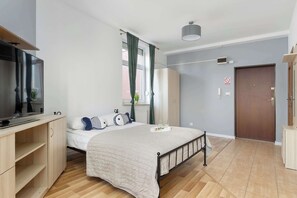 Ein Schlafzimmer mit Bett, Fernseher, Schreibtisch, Kleiderschrank, Tür und einem Fenster mit Vorhängen.