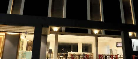 A building with a glass facade, a seating area with tables and chairs, and a staircase leading to the entrance.