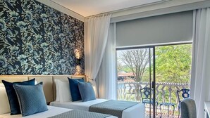 A hotel room with two beds, a patterned wall, a balcony with chairs, and a view of greenery. - Superior Twin room near the victoria falls (Livingstone)