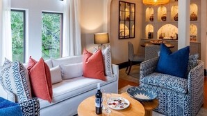 A living room with a white sofa, patterned armchairs, a round wooden table with a bottle and glasses, and a large woven light fixture. - Superior Twin room near the victoria falls (Livingstone)