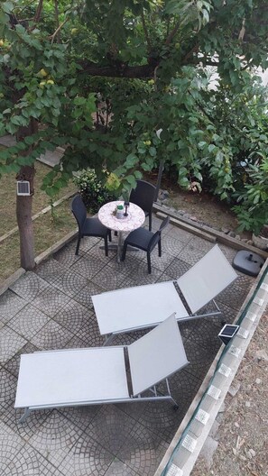 Un espacio al aire libre con una mesa redonda y sillas, rodeado por un jardín con árboles y un suelo empedrado.