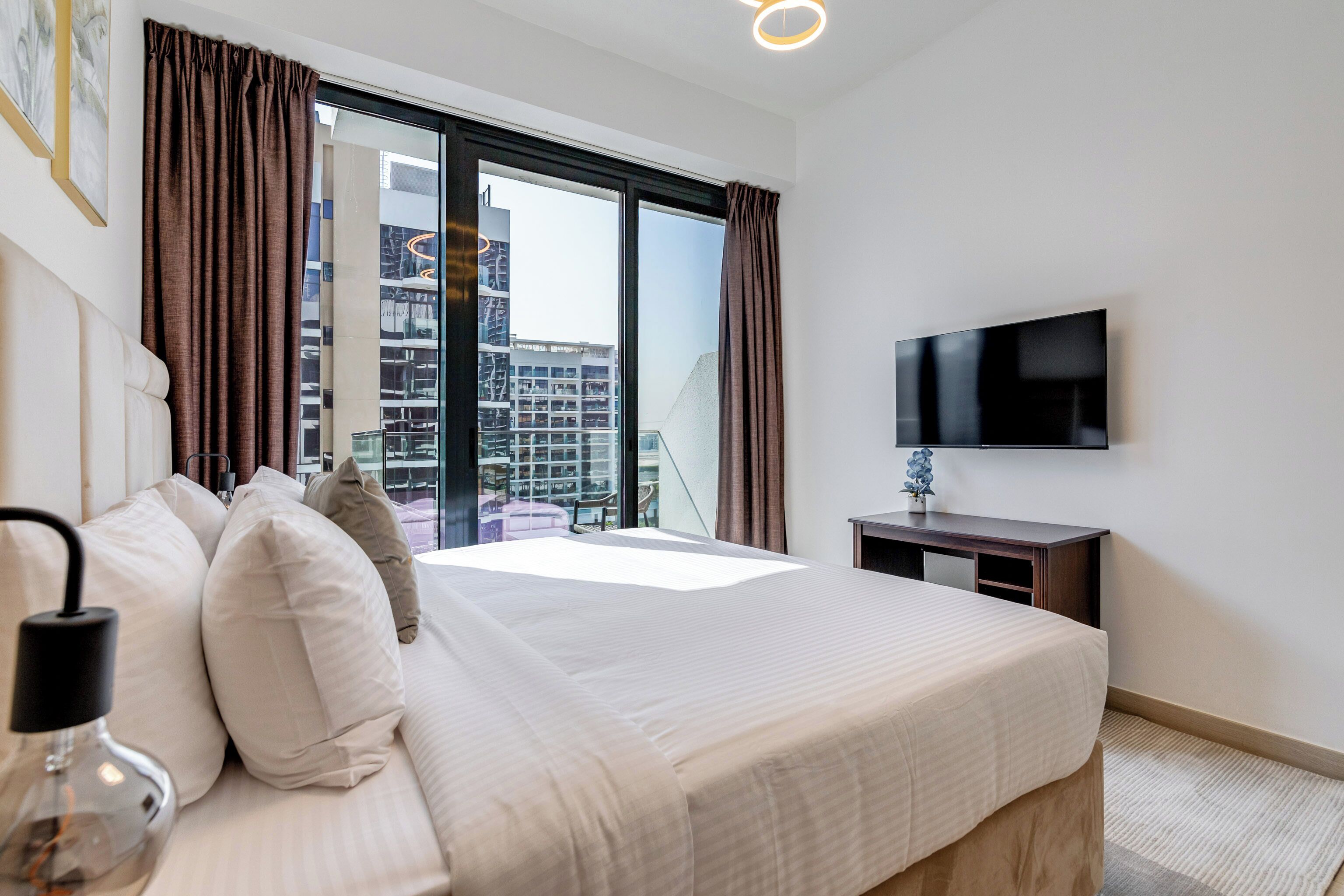 A hotel room with a large bed, a flat-screen TV, a desk, and a view of a cityscape through the window.