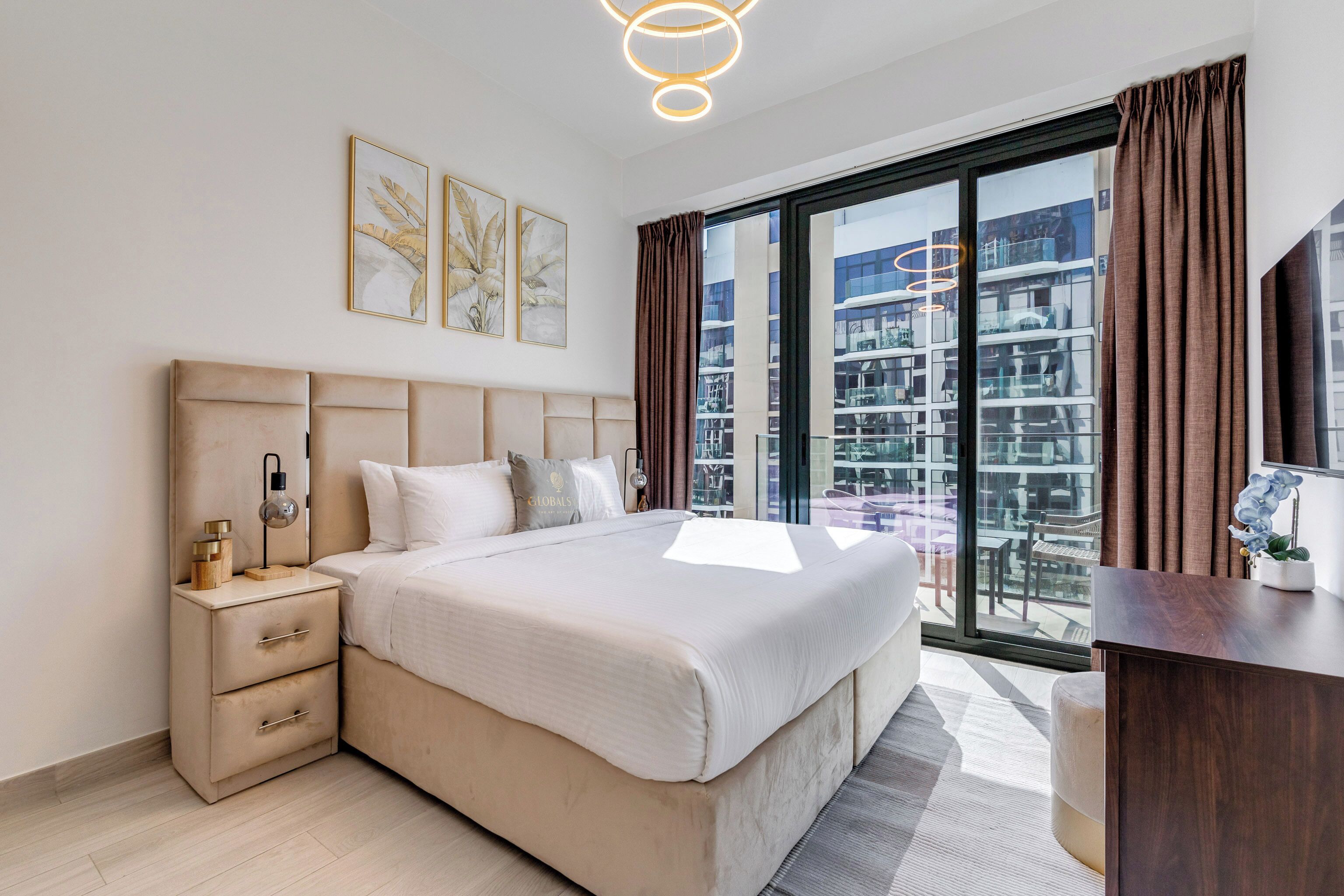 A modern bedroom with a large bed, a nightstand, a desk, and a view of a high-rise building.