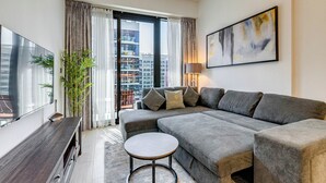 A modern living room with a grey sectional sofa, a round coffee table, a flat-screen TV, a potted plant, and a city view through large windows. - GlobalStay Azizi Riviera 42 (Dubai)