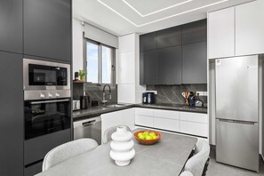 A modern kitchen with stainless steel appliances, a dining area with a table and chairs, and a bowl of apples. - The Emblem Residence by Nomads (Paphos)