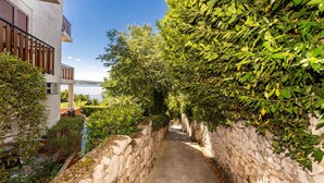 A narrow stone pathway flanked by lush greenery and a building with a balcony overlooking a body of water. - Beachfront apartment 1 - Dramalj by Villas Guide (Crikvenica)
