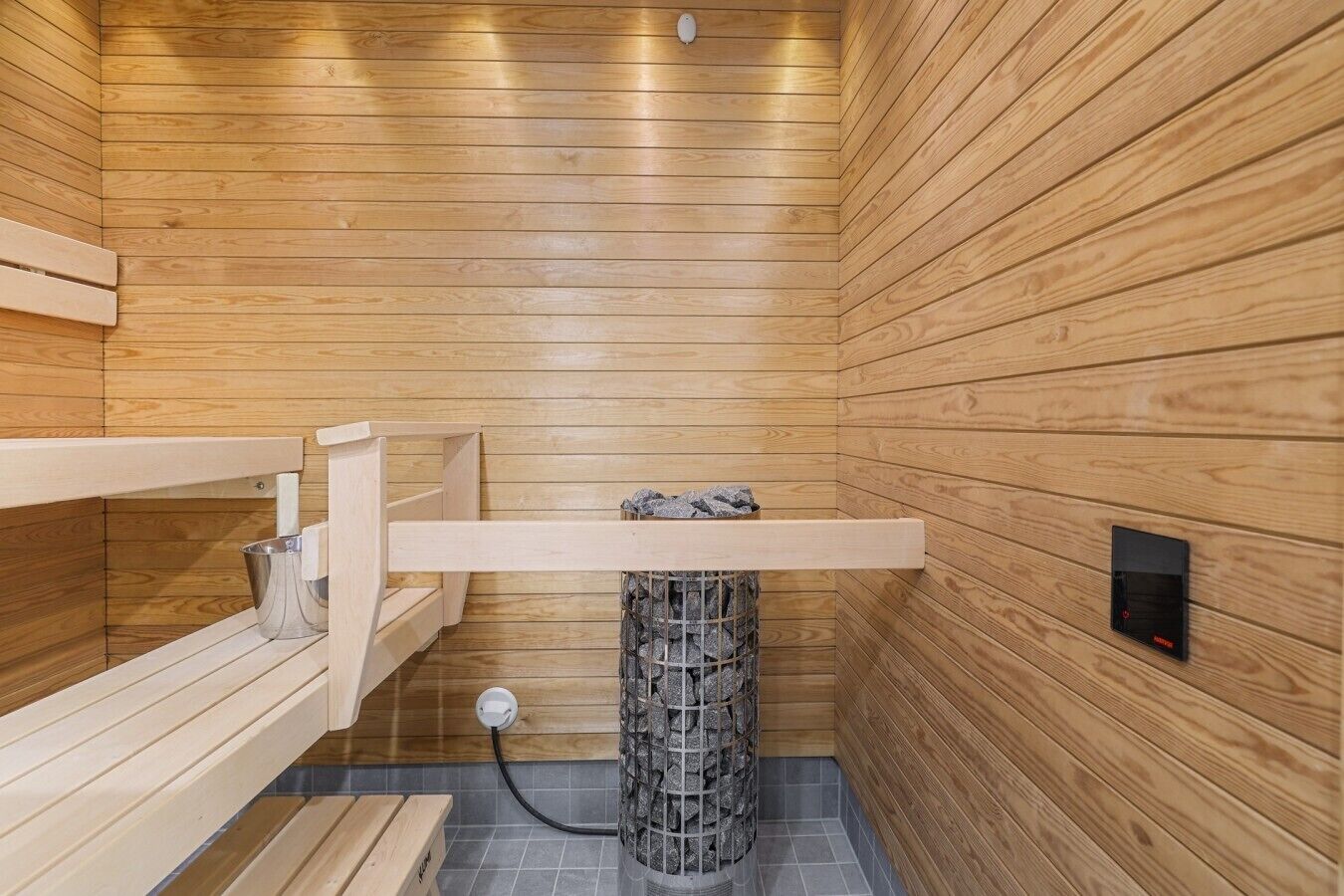 Una sala de sauna con paredes de madera, un banco, un calentador de piedra y un panel de control.