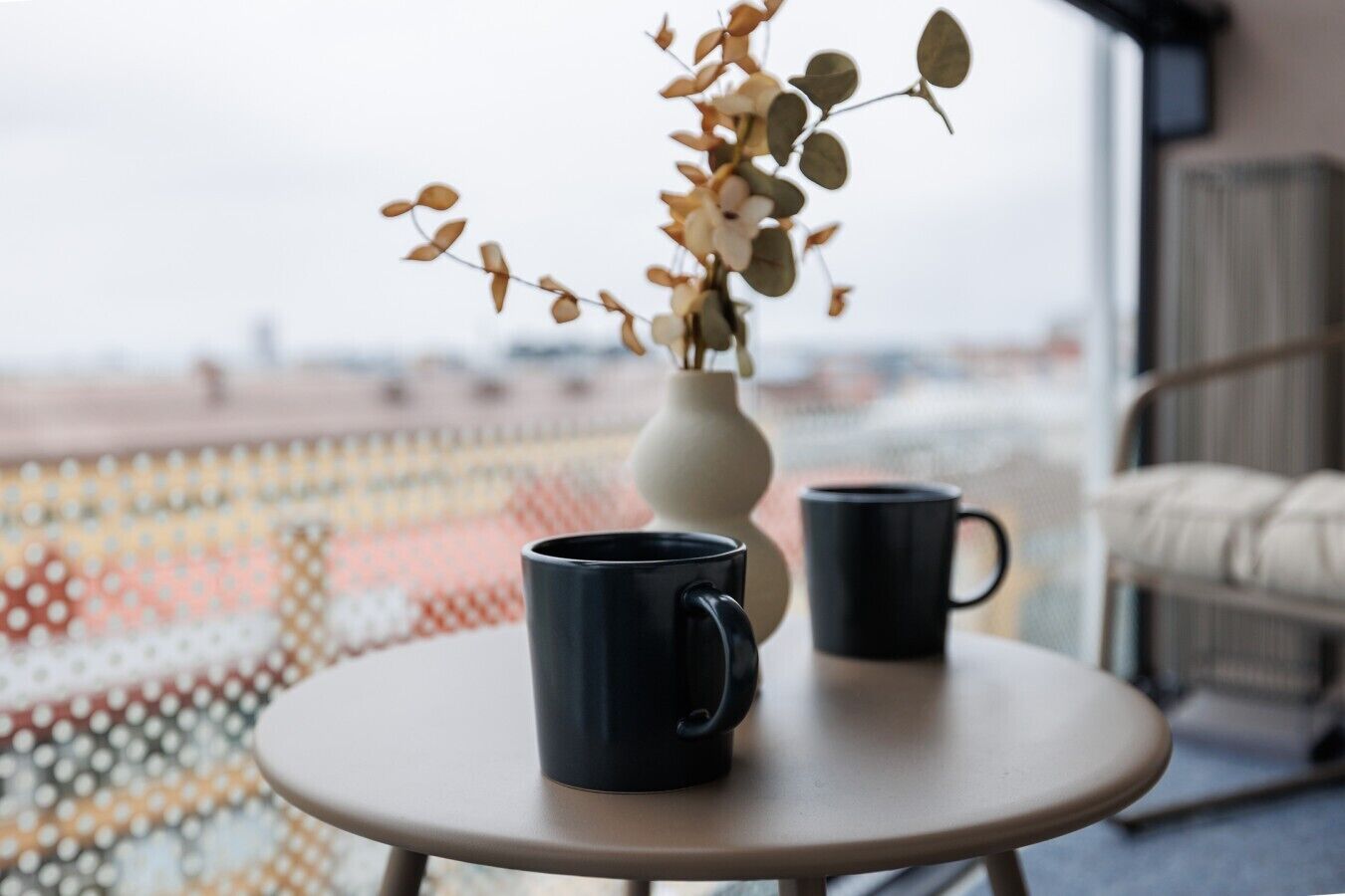 A round table with two black mugs and a white vase with dried flowers.