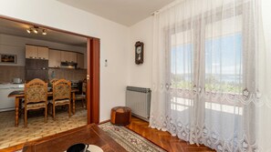 Ein Essbereich mit Holztisch und -stühlen, im Hintergrund eine Küche, ein Heizkörper und ein Fenster mit Spitzenvorhängen.