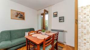 Ein gemütliches Wohnzimmer mit einem grünen Sofa, einem Holzesstisch und Stühlen. Ein Heizkörper, ein Fenster mit Vorhängen und gerahmte Bilder sind an der Wand zu sehen.