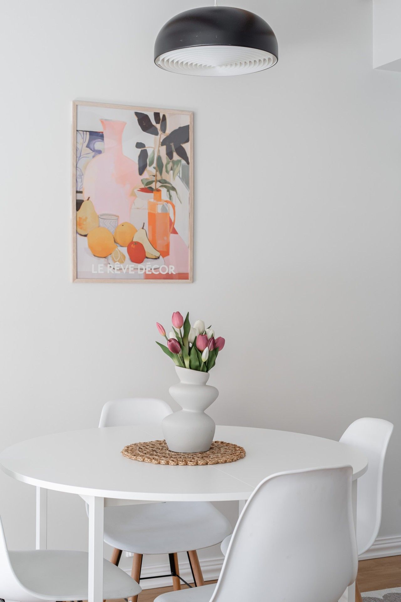 Ein weißer Esstisch mit einer weißen Blumenvase, umgeben von weißen Stühlen, unter einer schwarzen Pendelleuchte.
