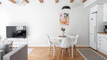 A modern living room with a flat-screen TV, a grey sofa, a dining table with white chairs, and a kitchen area with a white refrigerator.