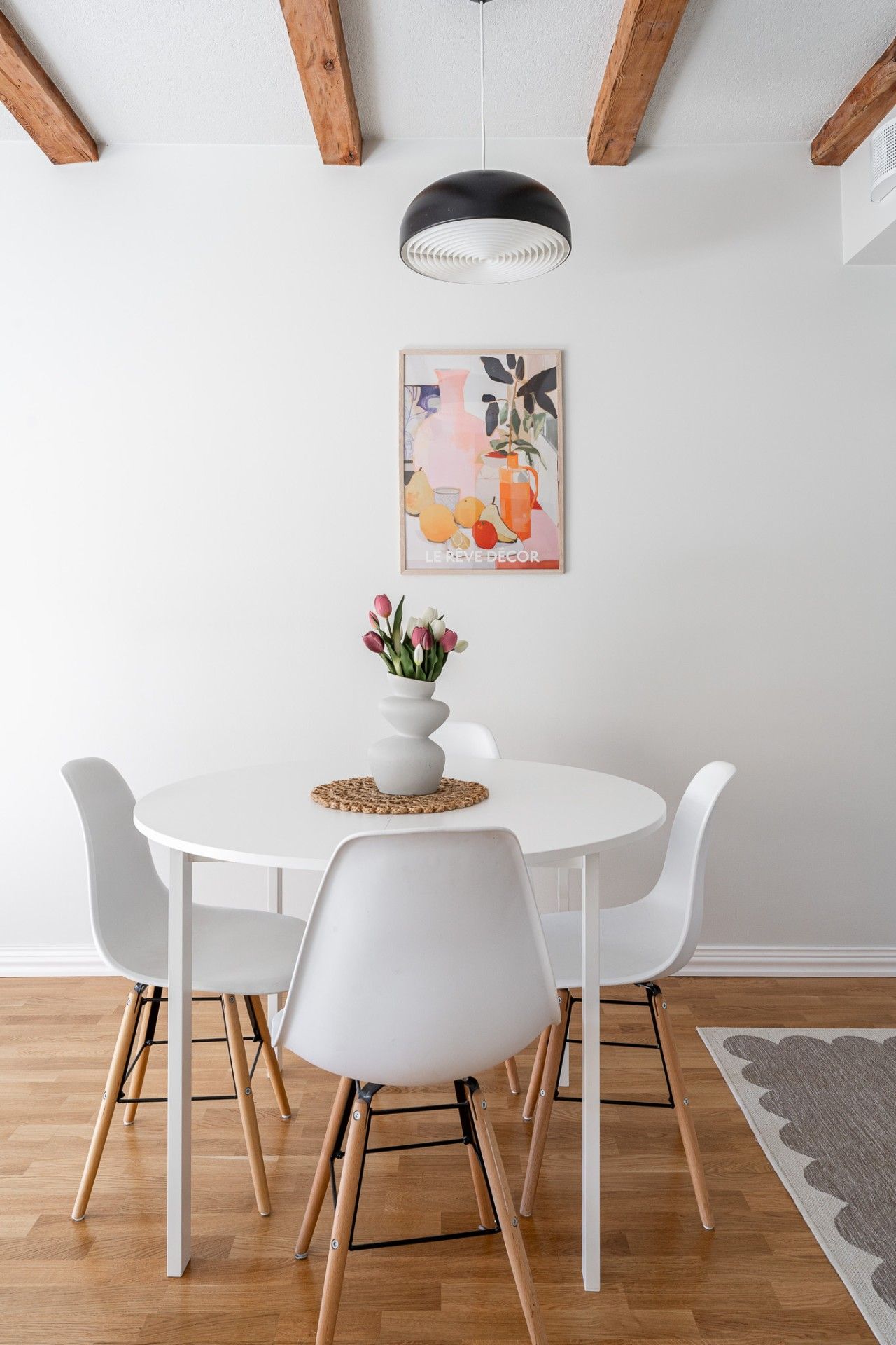 A dining area with a white table, four chairs, a vase with flowers, a hanging light fixture, and a framed artwork on the wall.