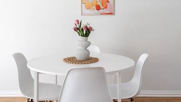 A dining area with a white table, four chairs, a vase with flowers, a hanging light fixture, and a framed artwork on the wall.