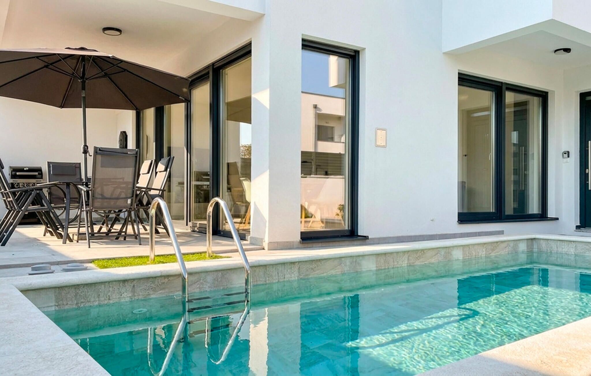 Een modern buitenzwembad met een ligbed en parasol, een eettafel met stoelen en een glazen deur naar een overdekt terras.