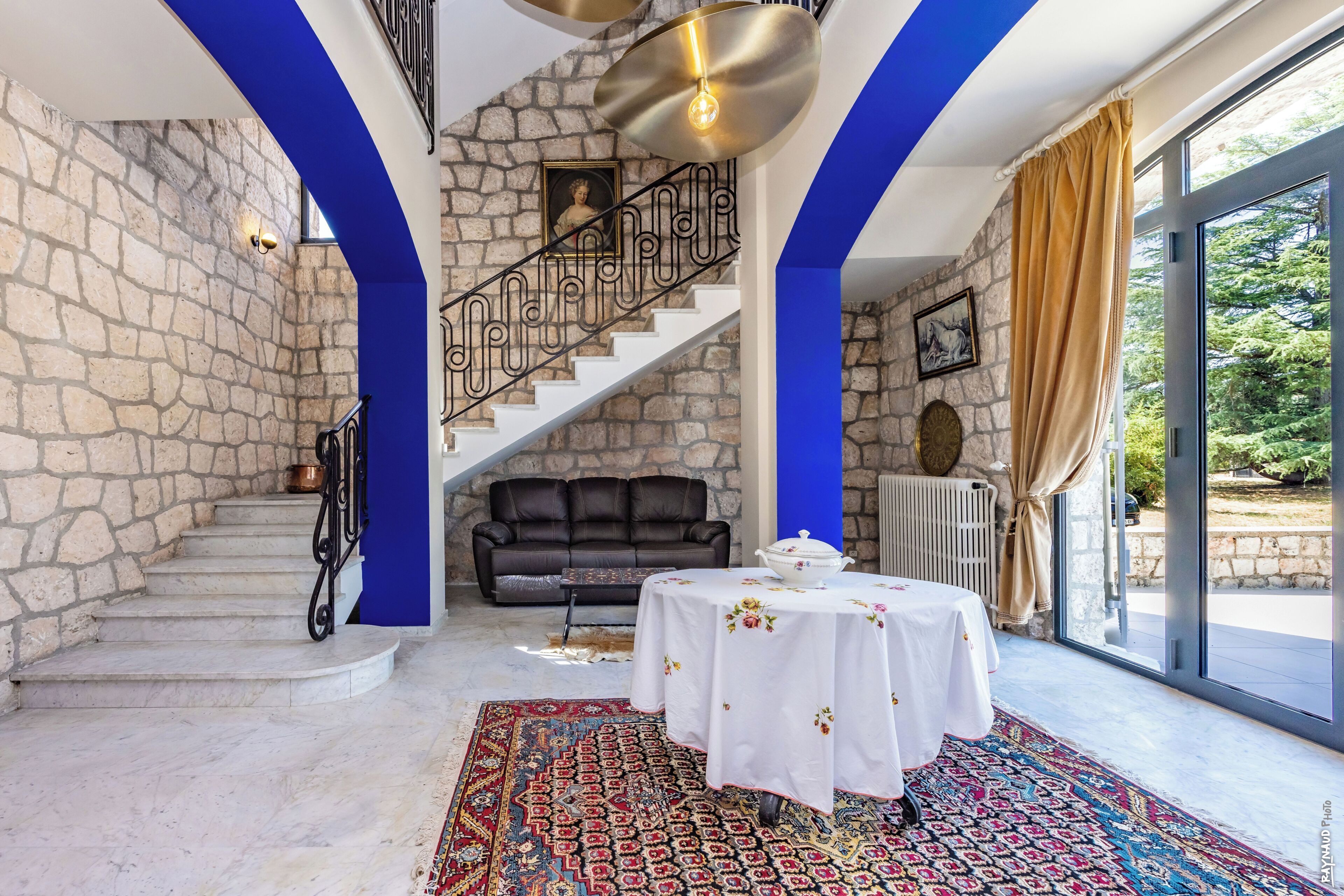 Une pièce avec un mur en pierre, un escalier, un canapé en cuir noir, une table ronde recouverte d’une nappe blanche et un tapis à motifs.
