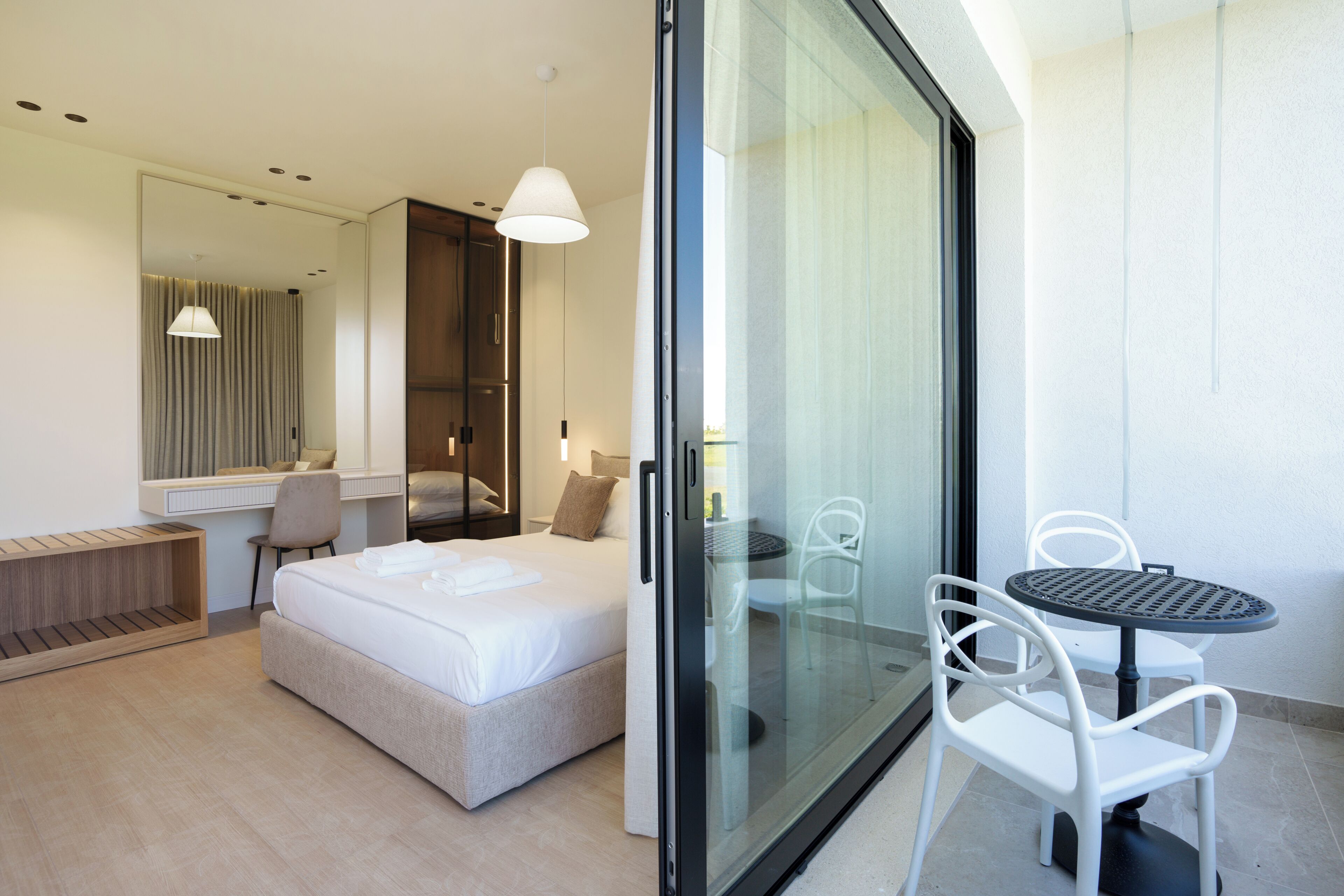 A modern hotel room with a bed, a desk, a chair, a table, and a sliding glass door leading to a balcony.
