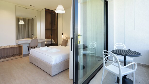 Ein modernes Hotelzimmer mit Bett, Schreibtisch, Stuhl, Tisch und einer Schiebetür, die auf einen Balkon führt.