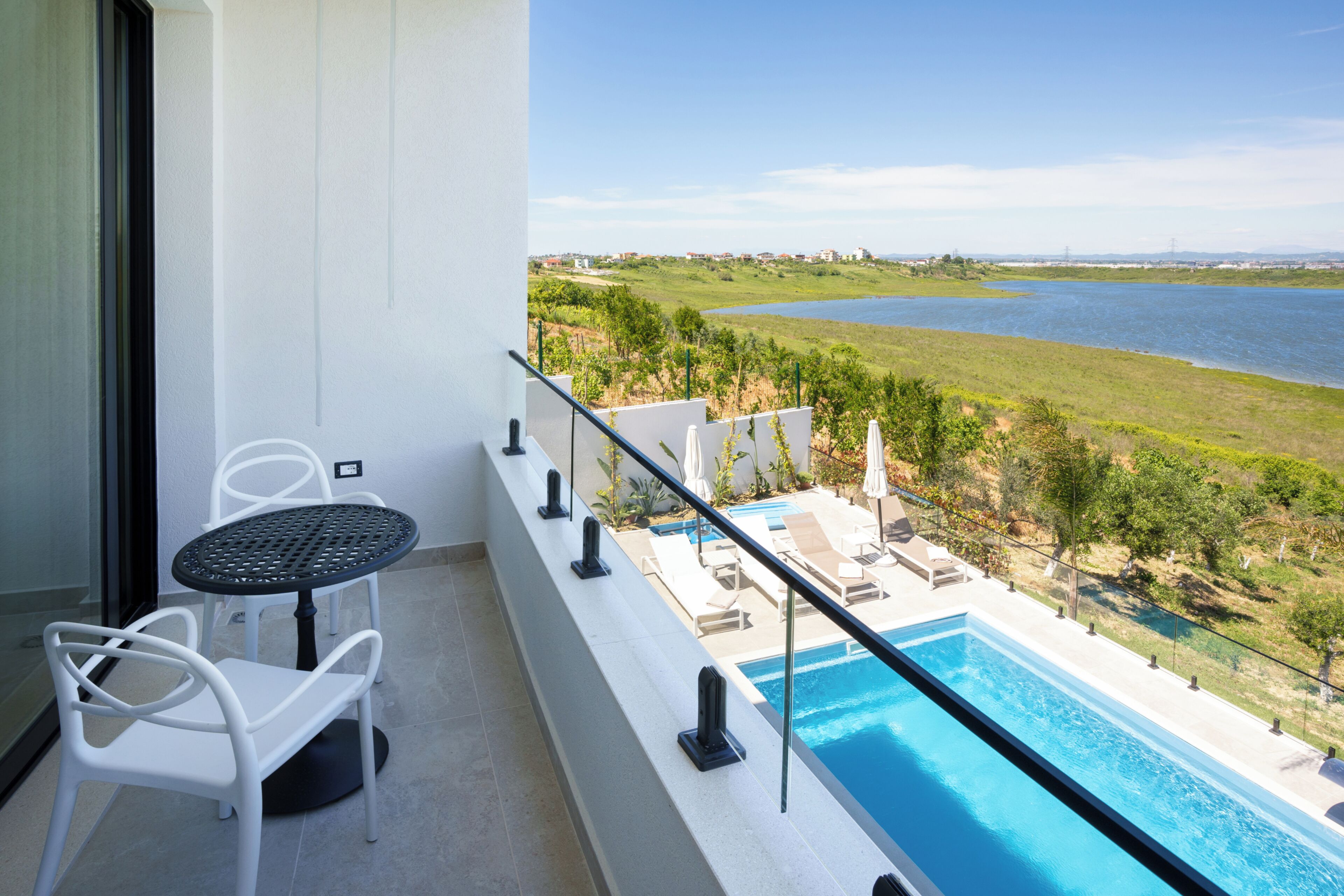 A balcony with a pool, white chairs, and a view of a lake and distant buildings.