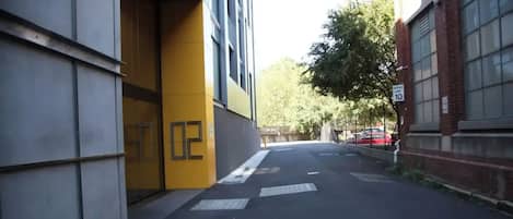 Eine schmale Stadtstraße mit einem Tempolimit-Schild, einem gelben Gebäude und einem roten Auto, das am Straßenrand geparkt ist.