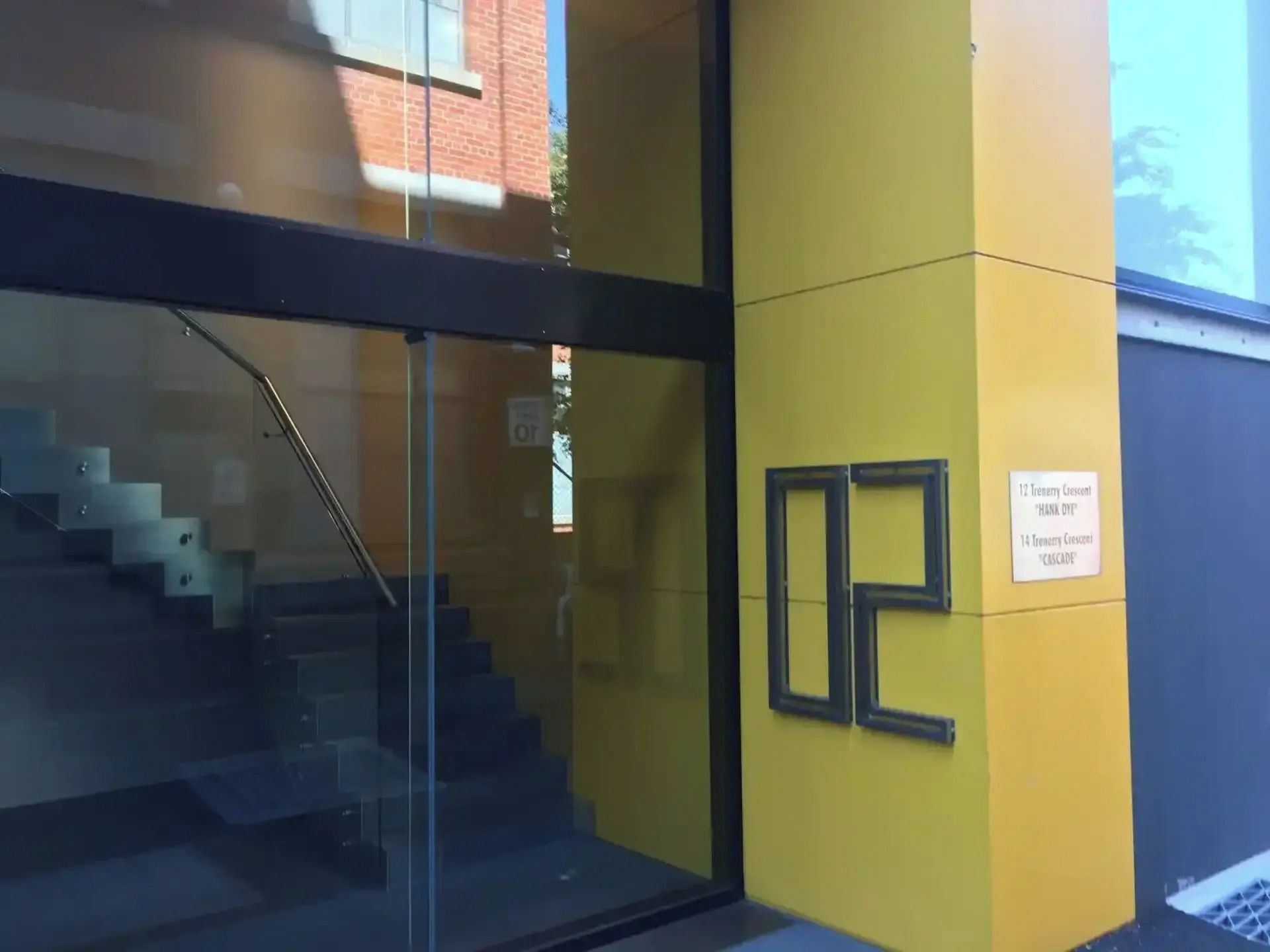 A building entrance with a glass door, a staircase, and a sign indicating '12 Trenchy Crescent 'Thank You''.