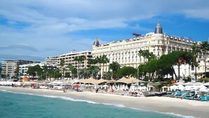 Ein Strandhotel mit mehreren Stockwerken, einer markanten Kuppel und einer Strandlage mit Sonnenschirmen und Liegestühlen.