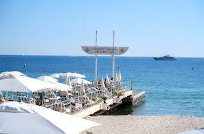 A beachside lounge with umbrellas and lounge chairs, a sign indicating 'Carlton Cannes The Beach Club,' and a yacht in the background. - Apartment 'D&L Sea View' with Private Terrace, Wi-Fi and Air Conditioning (Cannes)