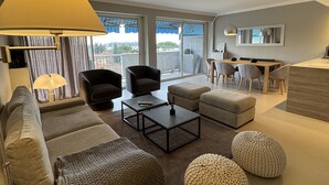 Ein modernes Wohnzimmer mit einer Couch, Sesseln, Hocker, einem Couchtisch und einem Essbereich mit Stühlen und einem Tisch.
