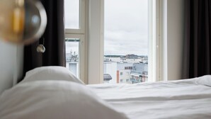 A bed with white linens, a view of buildings through a window, and a hanging light fixture. - New 2BR Apt with Sauna & Stunning Views  (Oulu)