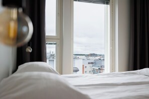 Una cama con ropa blanca, vista de edificios a través de una ventana y una lámpara colgante.