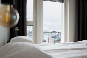Una cama con ropa blanca, vista de edificios a través de una ventana y una lámpara colgante.