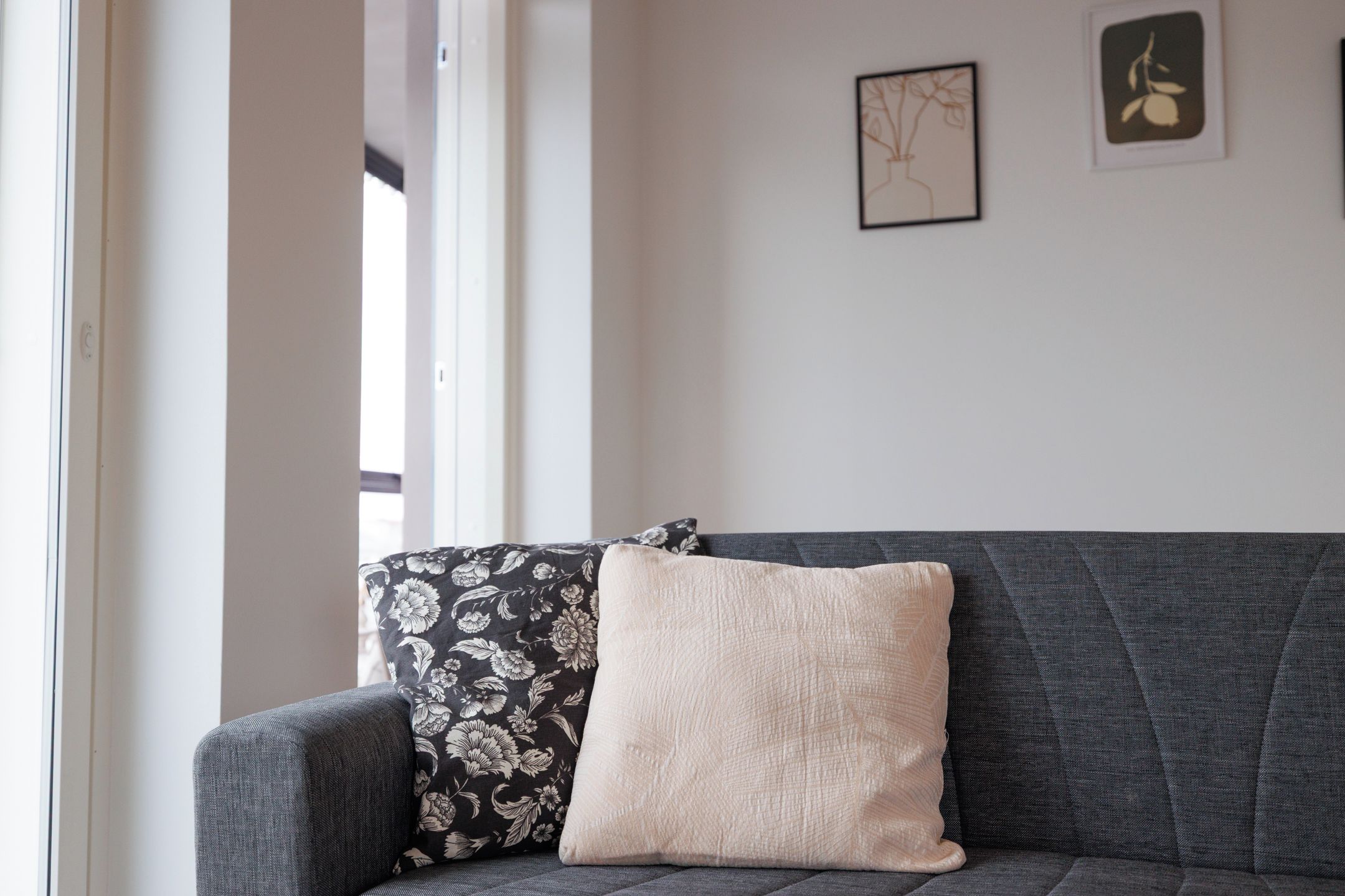 A sofa with two pillows, one with a floral pattern and one plain, in a room with a plain wall and a window.