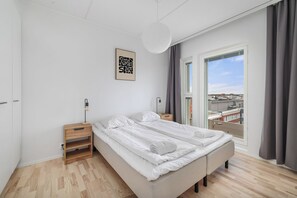A modern bedroom with a large bed, bedside tables, a hanging light fixture, and a view of the city through a large window. - New 2BR Apt with Sauna & Stunning Views  (Oulu)